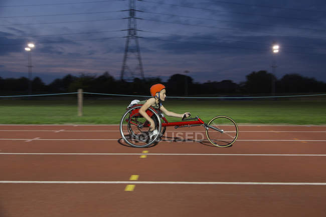 Adolescente chica parapléjico atleta en silla de ruedas carrera en pista deportiva por la noche - foto de stock