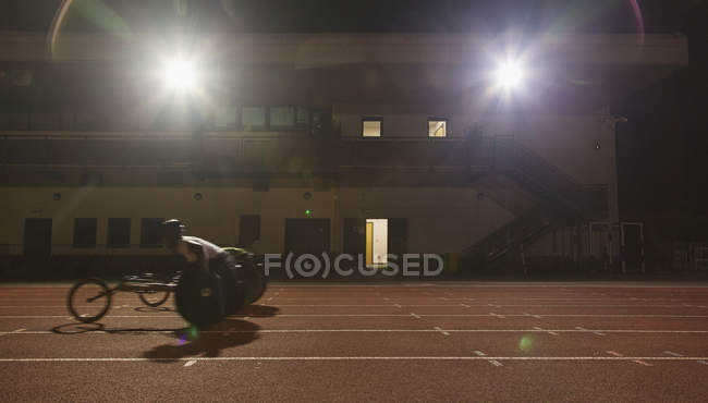 Entrenamiento de atleta parapléjico masculino para la carrera en silla de ruedas en pista deportiva por la noche - foto de stock