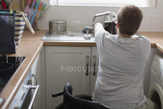 Jovem mulher em cadeira de rodas enchendo chaleira para chá na pia da cozinha do apartamento — Fotografia de Stock