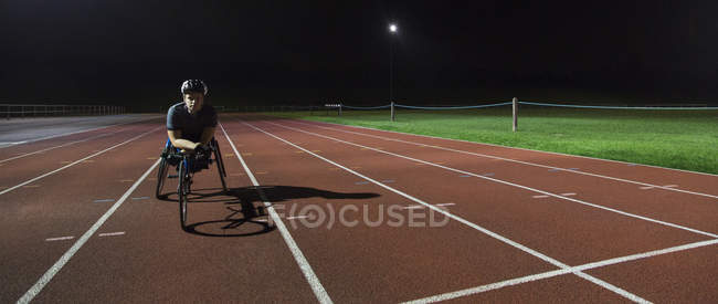 Retrato seguro, determinado joven atleta parapléjico entrenamiento para la carrera en silla de ruedas en pista deportiva por la noche - foto de stock