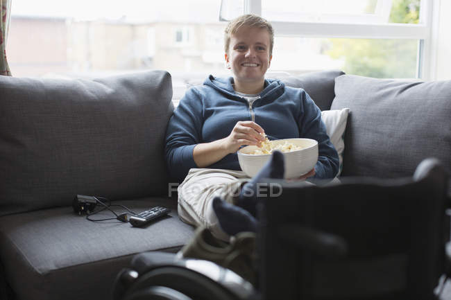 Jeune femme souriante regardant la télévision et mangeant du pop-corn sur un canapé avec les pieds levés sur un fauteuil roulant — Photo de stock