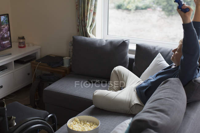 Mujer joven animando, jugando videojuegos en el sofá junto a la silla de ruedas - foto de stock