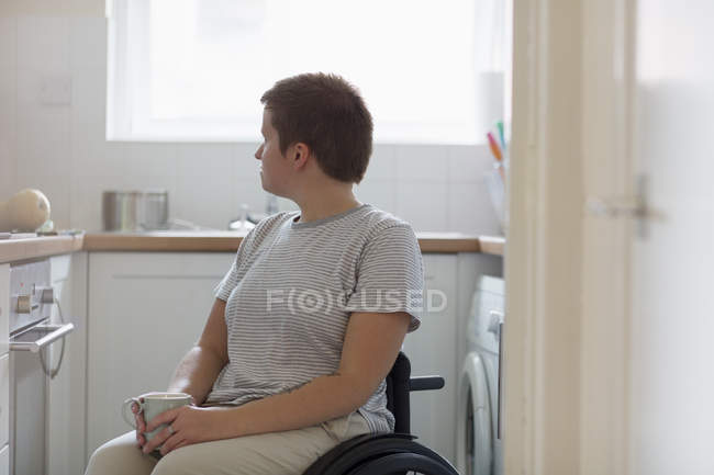 Thoughtful young woman in wheelchair drinking tea in apartment kitchen — Stock Photo