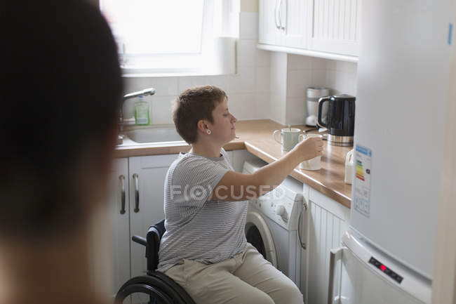 Jovem mulher em cadeira de rodas preparando chá na cozinha do apartamento — Fotografia de Stock