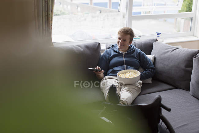 Young woman watching TV and eating popcorn on sofa with feet up on wheelchair — Stock Photo
