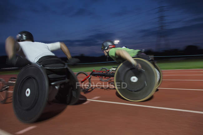 Atletas paraplégicos acelerando ao longo da pista de esportes durante a corrida em cadeira de rodas à noite — Fotografia de Stock