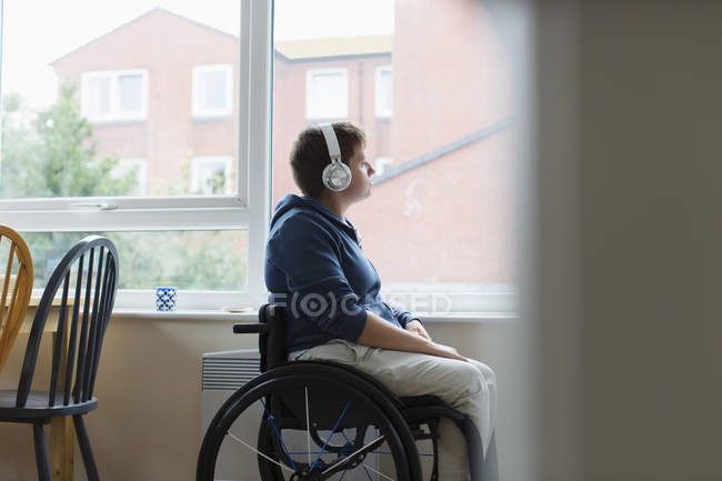 Nachdenkliche junge Frau im Rollstuhl hört Musik mit Kopfhörern am Fenster — Stockfoto