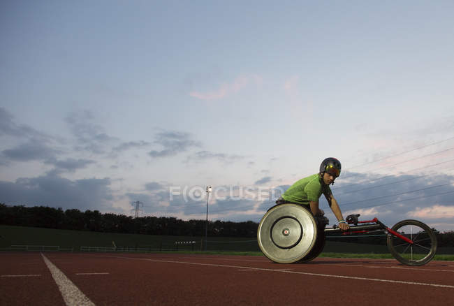 Joven atleta parapléjico masculino entrenando para la carrera en silla de ruedas en pista deportiva por la noche - foto de stock