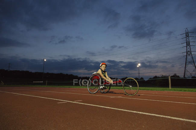 Retrato confiante adolescente menina paraplégica atleta treinamento para corrida em cadeira de rodas na pista de esportes à noite — Fotografia de Stock
