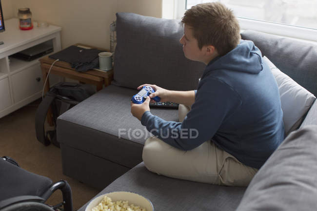 Молодая женщина играет в видеоигры на диване рядом с инвалидной коляской — стоковое фото