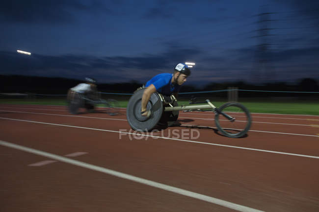 Decidido joven atleta parapléjico masculino exceso de velocidad a lo largo de la pista deportiva en la carrera en silla de ruedas por la noche - foto de stock