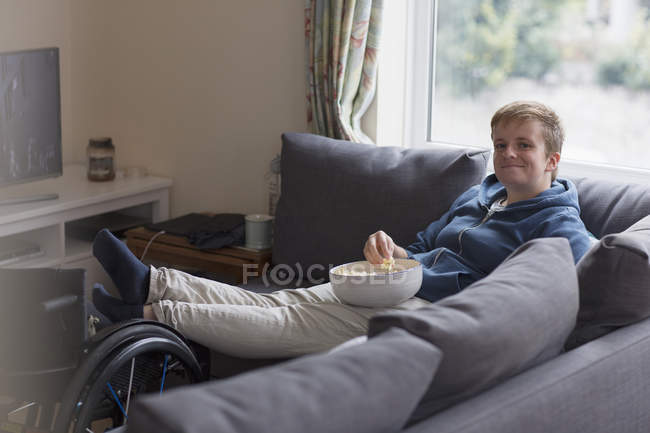 Уверенная в себе молодая женщина смотрит телевизор и ест попкорн на диване с ногами на инвалидной коляске — стоковое фото