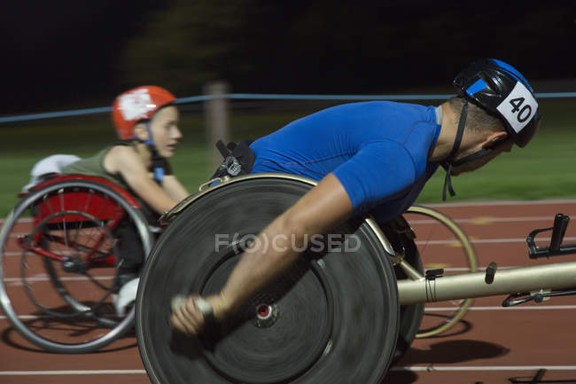 Atletas paraplégicos acelerando ao longo da pista de esportes em corrida de cadeira de rodas à noite — Fotografia de Stock