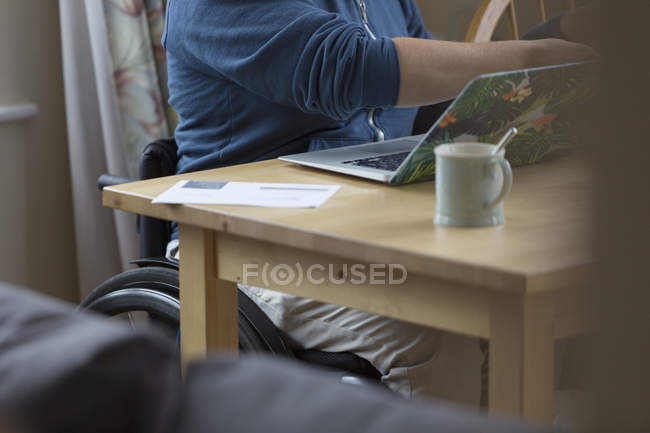 Jovem mulher em cadeira de rodas usando laptop na mesa de jantar — Fotografia de Stock