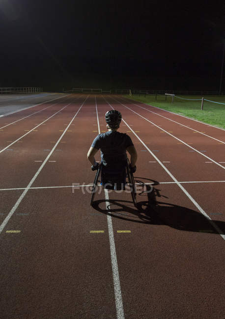 Decidida joven atleta parapléjica entrenamiento para la carrera en silla de ruedas en pista deportiva por la noche - foto de stock