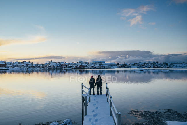 Coppia che si tiene per mano ai margini del bacino innevato con vista sul villaggio lungomare, Reine, Isole Lofoten, Norvegia — Foto stock