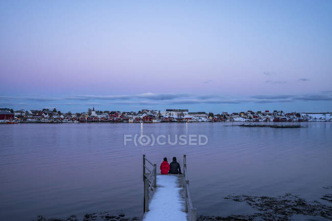 Couple sitting at the edge of snowy pier overlooking waterfront fishing village, Reine, Lofoten Islands, Norway — Stock Photo