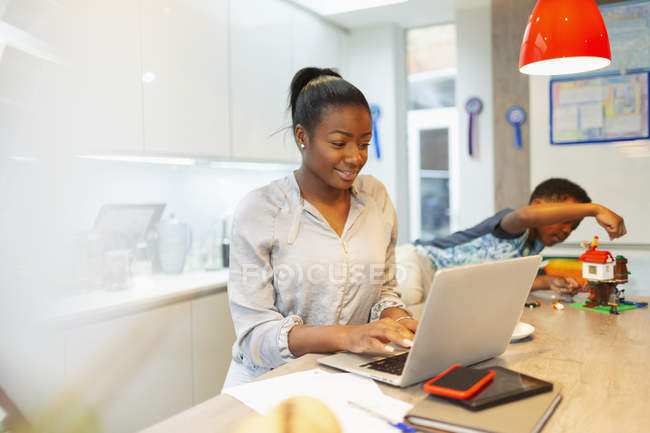 Boy playing next to mother working at laptop in kitchen — Stock Photo
