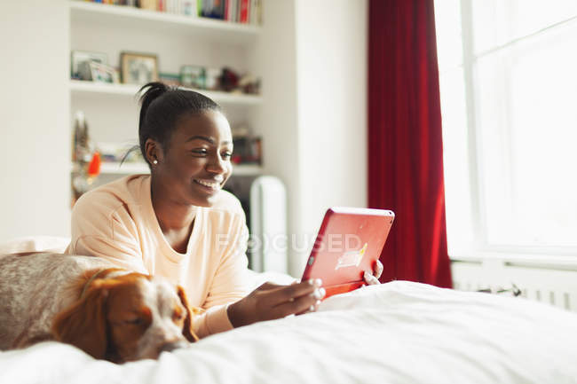 Sorrindo jovem mulher usando tablet digital ao lado de cão adormecido na cama — Fotografia de Stock
