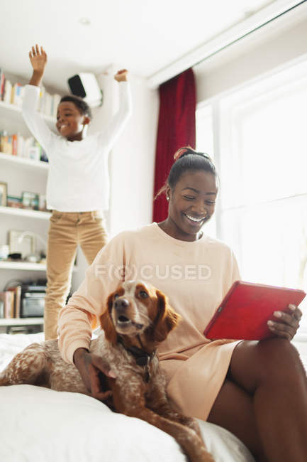 Rapaz brincalhão pulando na cama atrás de cão e mãe com tablet digital — Fotografia de Stock