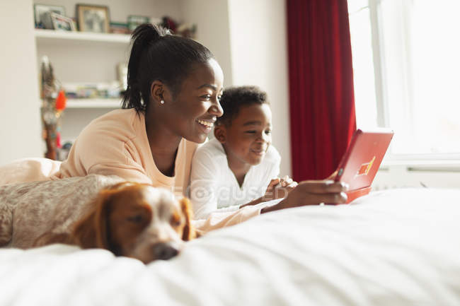 Madre e hijo usando tableta digital en la cama al lado del perro dormido - foto de stock