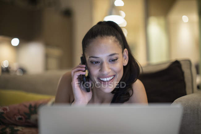 Усміхнена жінка говорить на смартфоні на ноутбуці на дивані — стокове фото