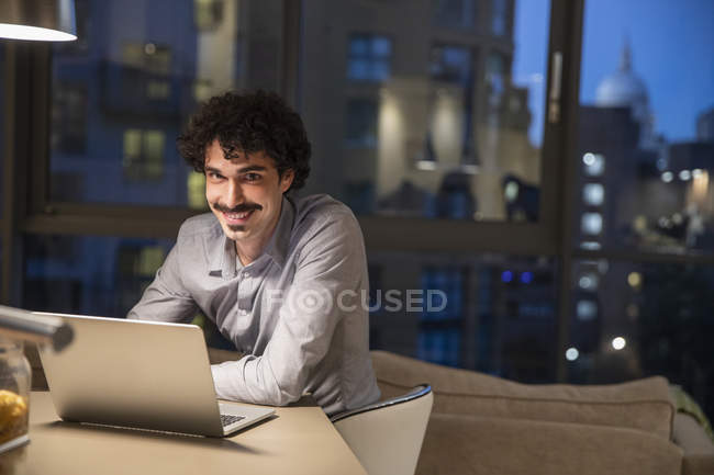 Retrato sorrindo homem usando laptop em apartamento urbano à noite — Fotografia de Stock