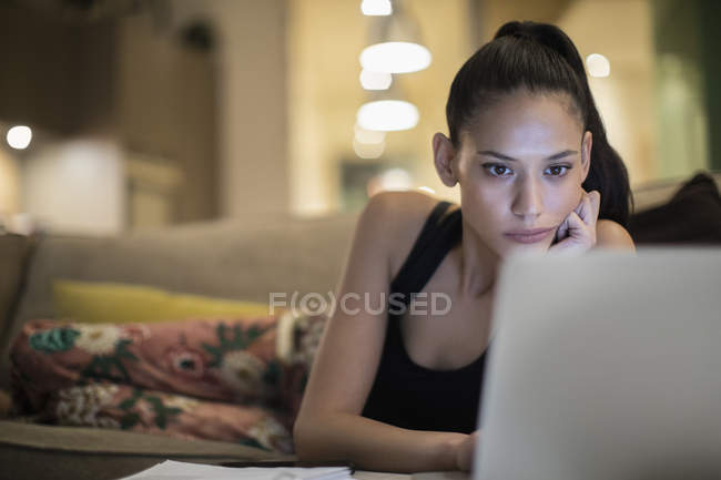 Зосереджена жінка в піжамі, використовуючи ноутбук на дивані — стокове фото