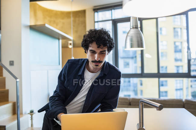 Hombre enfocado que trabaja en el ordenador portátil en la cocina del apartamento - foto de stock