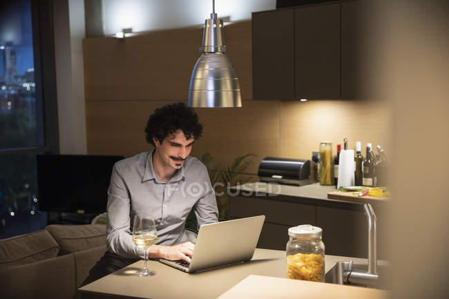 Homem bebendo vinho branco no laptop na cozinha do apartamento à noite — Fotografia de Stock