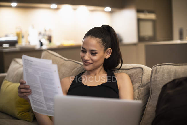 Усміхнена жінка читає документи та використовує ноутбук на дивані — стокове фото