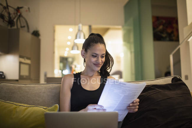 Mujer leyendo papeleo en el portátil en el sofá de la sala de estar - foto de stock