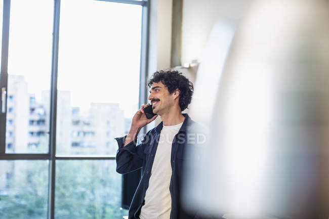 Усміхнений чоловік розмовляє по смартфону у вікні міської квартири — стокове фото
