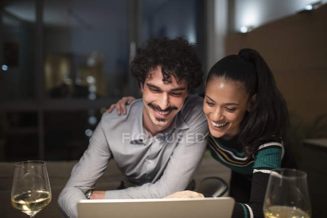 Couple heureux utilisant un ordinateur portable et boire du vin blanc à la maison la nuit — Photo de stock