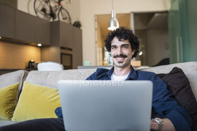 Hombre sonriente retrato usando portátil en sofá de apartamento - foto de stock