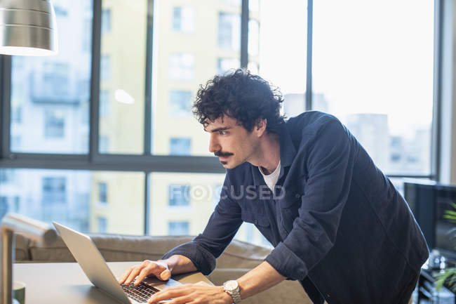 Hombre enfocado utilizando el ordenador portátil en la cocina apartamento urbano - foto de stock