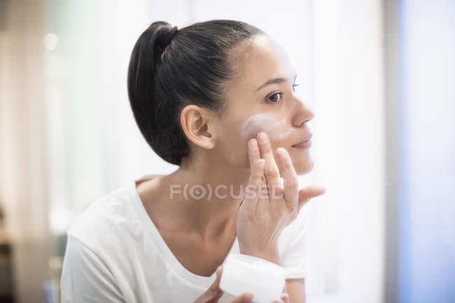 Woman applying moisturizer to face in bathroom mirror — Stock Photo