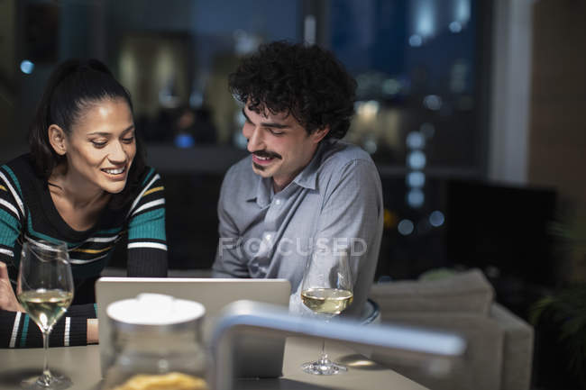 Smiling couple using laptop and drinking white wine at home at night — Stock Photo