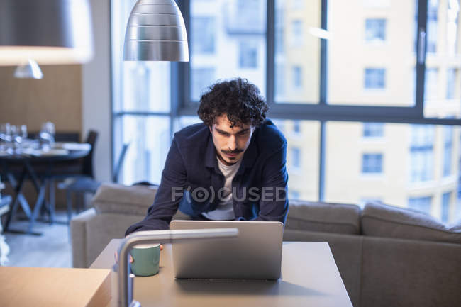 Man with coffee working at laptop in apartment kitchen — Stock Photo