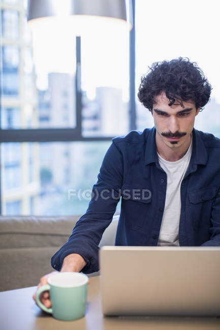 Человек с кофе работает за ноутбуком в квартире — стоковое фото