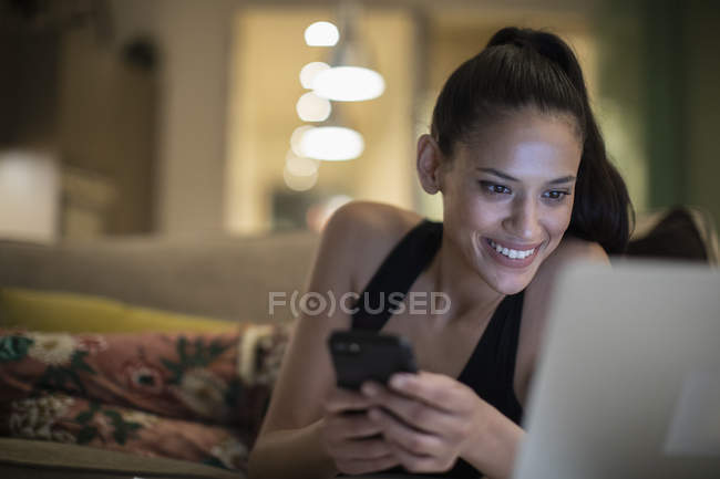 Усміхнена жінка в піжамі використовує смартфон і ноутбук на дивані — стокове фото