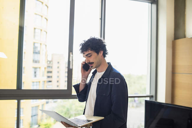 Hombre usando el ordenador portátil y hablando en el teléfono inteligente en la ventana del apartamento urbano - foto de stock