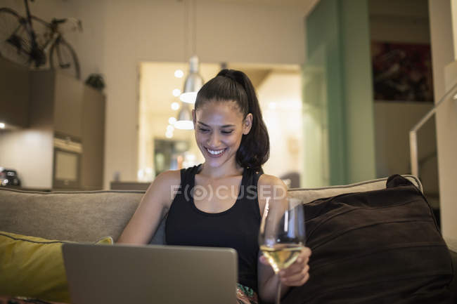 Mujer sonriente usando laptop y bebiendo vino blanco en el sofá del apartamento - foto de stock