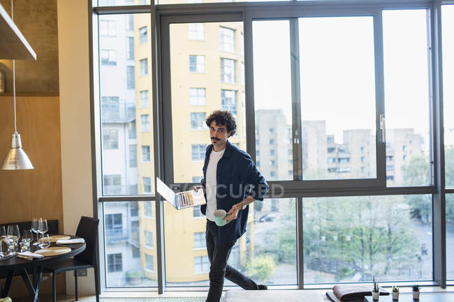 Hombre con portátil y café caminando en apartamento urbano - foto de stock
