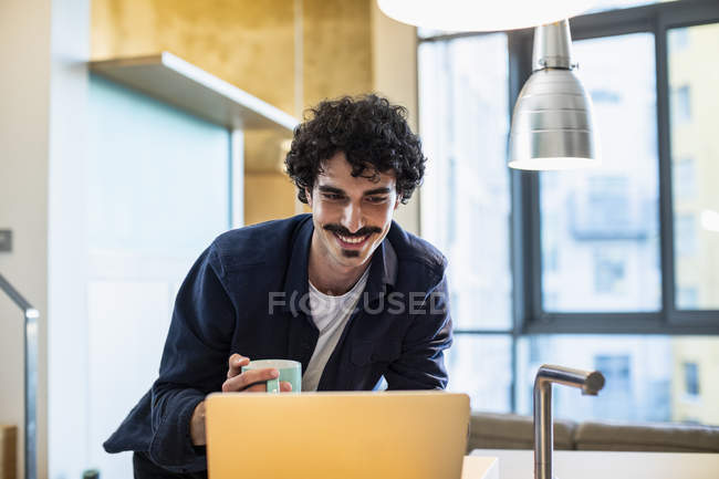 Улыбающийся мужчина пьет кофе, работает за ноутбуком на кухне — стоковое фото