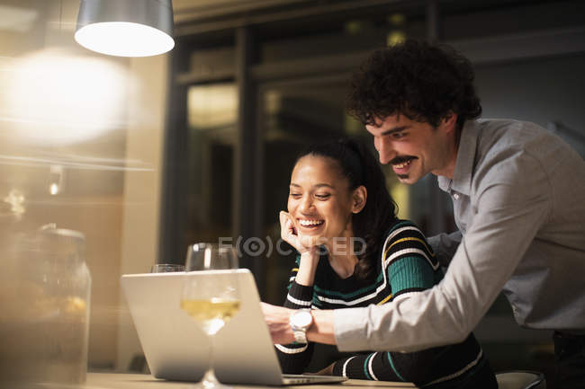 Pareja feliz usando el ordenador portátil y beber vino blanco en casa por la noche - foto de stock