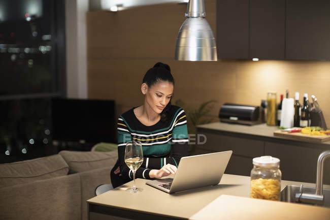 Fokussierte Frau benutzt Laptop und trinkt nachts Weißwein in Wohnküche — Stockfoto
