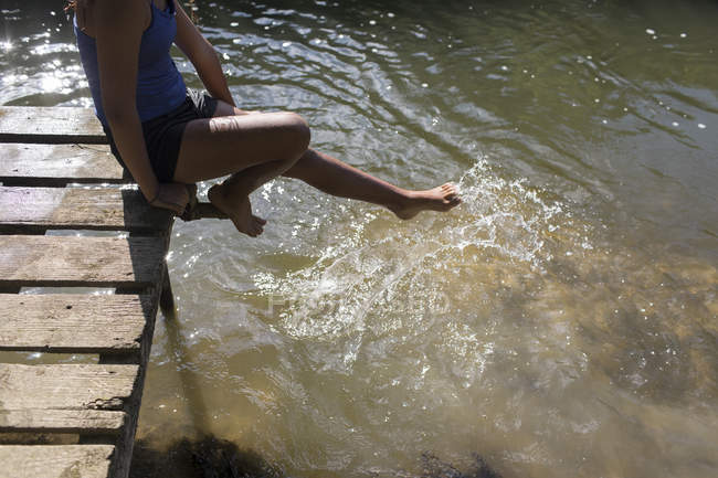 Menina na doca ensolarada espirrando pé na água do rio — Fotografia de Stock
