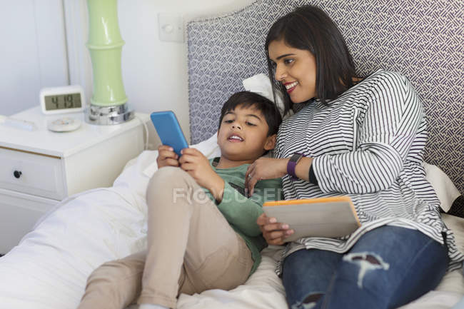 Madre e hijo usando teléfono inteligente y tableta digital en la cama - foto de stock