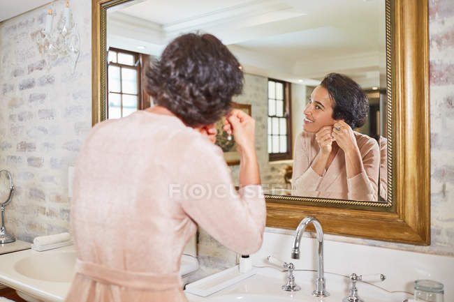 Mujer poniendo pendientes en el espejo del baño del hotel - foto de stock